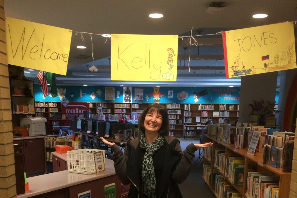 Author Kelly Jones (a white woman) smiles in a school library under a yellow banner that reads "Welcome Kelly Jones" with student art.